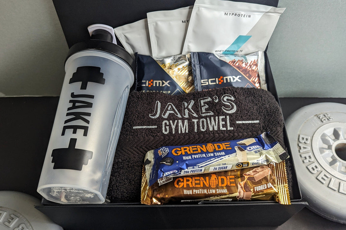 Open black fitness gift box containing a shaker, protein snacks, small protein packets and a personalized gym towel