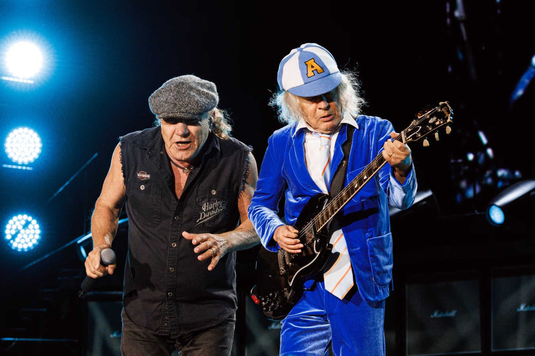AC/DC members Brian Johnson and Angus Young performing live, with Angus playing a black electric guitar in a blue stage outfit.