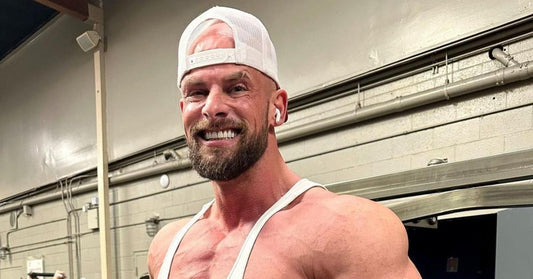 Joey Swoll smiling in a white tank top and backward cap inside a gym