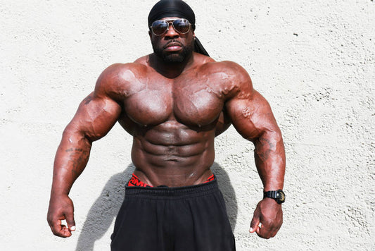 Kali Muscle shirtless in a dark gym wearing a black head covering and holding a heavy dumbbell in each hand, arms and chest flexed