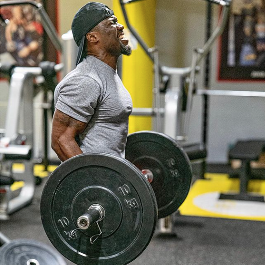 Kevin Hart performing a barbell deadlift in a gym while wearing a gray training shirt and backward cap