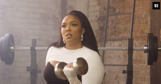 Lizzo performing a dumbbell curl in a gym wearing a white-and-black athletic outfit