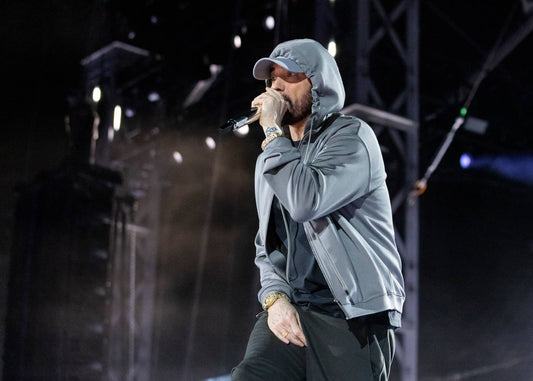 Eminem performing into a microphone onstage in a gray hooded sweatshirt and cap during a Detroit concert.