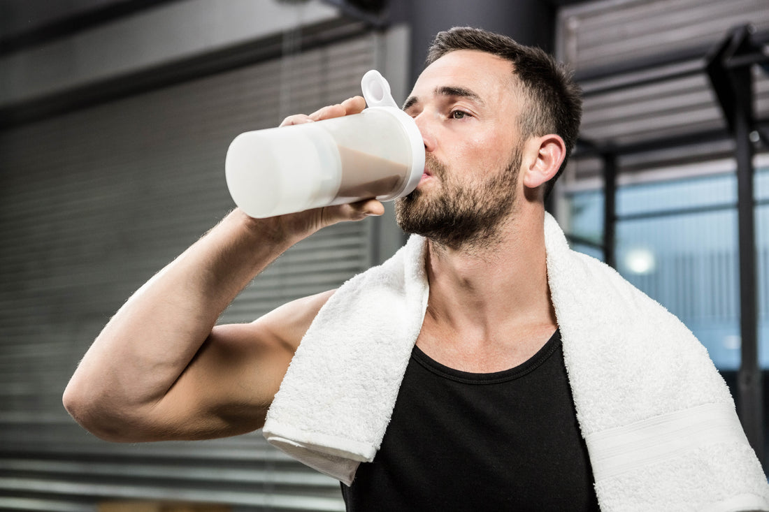 Muscular man in a black tank top with a towel over his shoulders drinking a white protein shaker after training