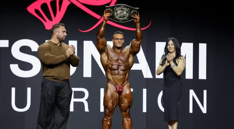 Nick Walker celebrating his 2026 Mr. Olympia victory onstage beside the event host and prize display.