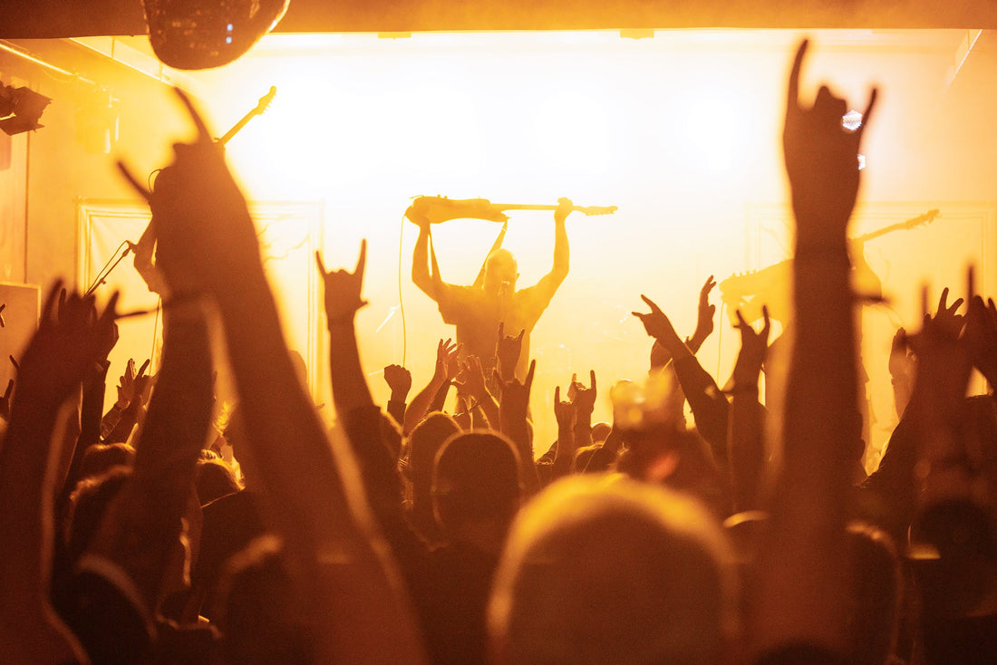 Live metal guitarist holding a guitar above a crowd with raised hands under golden stage lighting