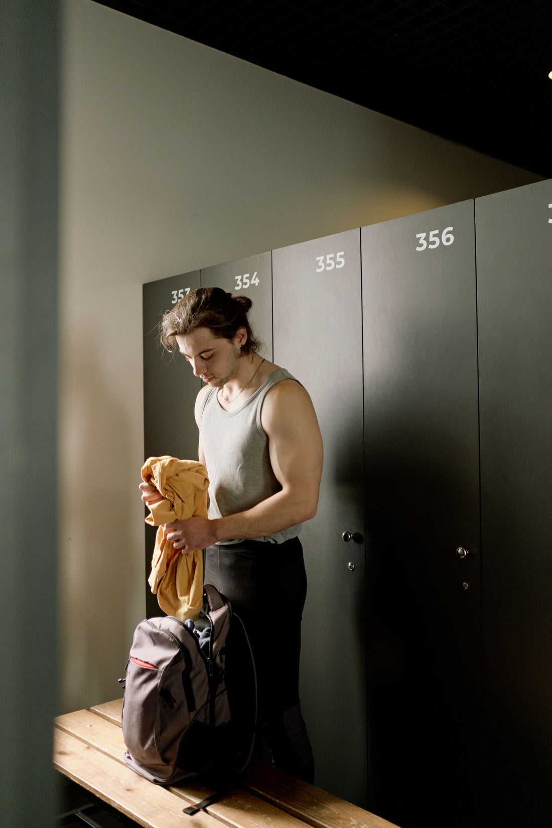 Man in a gray sleeveless top sorting gym clothing into a backpack in front of numbered locker doors
