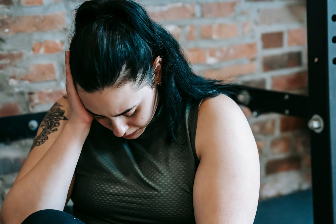 Woman sitting on a gym floor near exercise equipment, looking tired and stressed