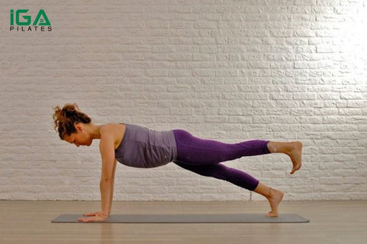 Woman performing a Pilates leg pull front in a high plank with one straight leg extended behind her on a studio mat.