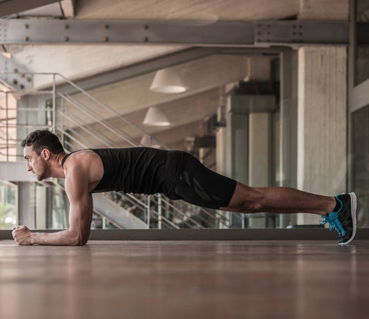 Man in black workout clothing holding a straight-body forearm plank on a wooden gym floor