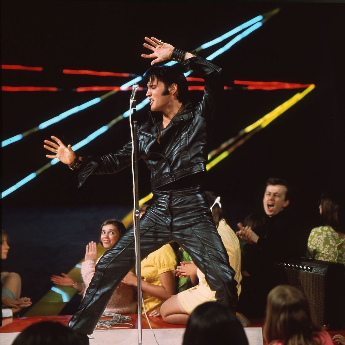 Elvis Presley performing in black leather at a Las Vegas show with a microphone and bright stage lights