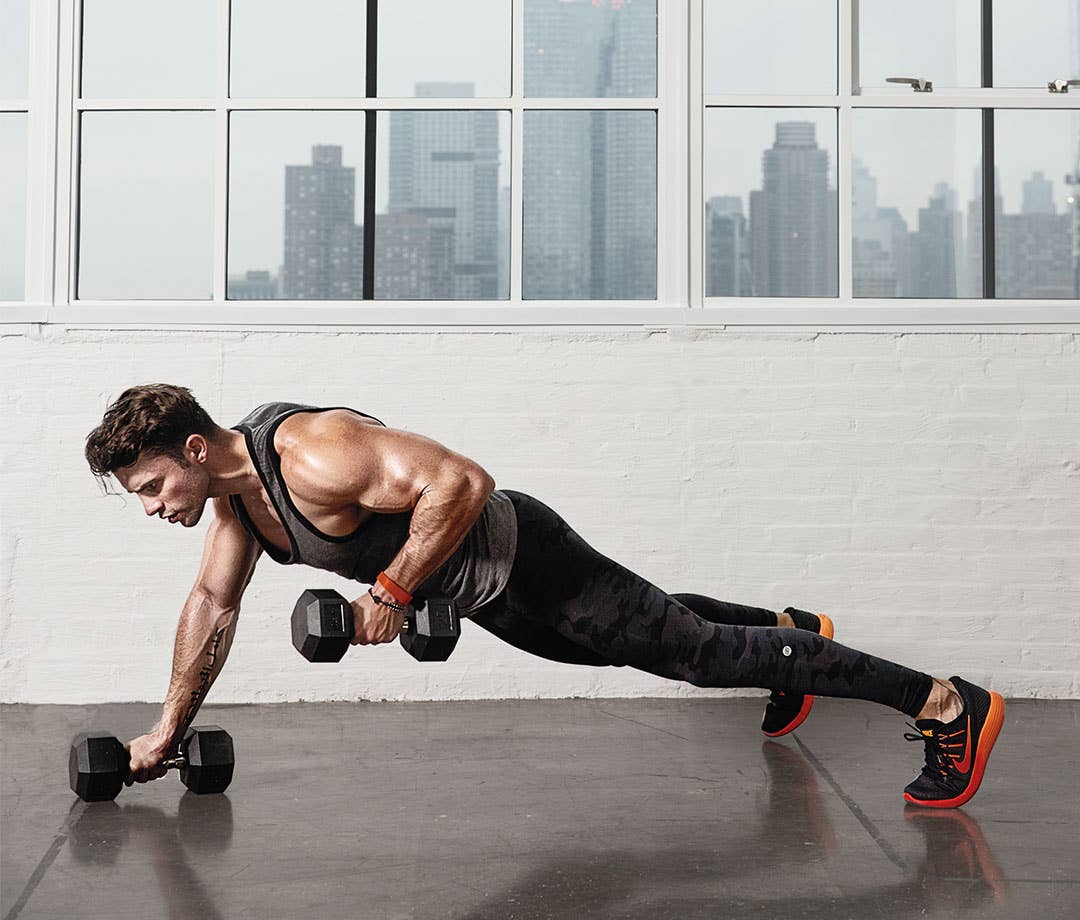 Man performing a renegade dumbbell row in a high plank in a bright gym