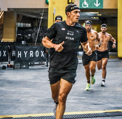 Rich Ryan running at the front of a HYROX indoor race wearing a black RYAN jersey