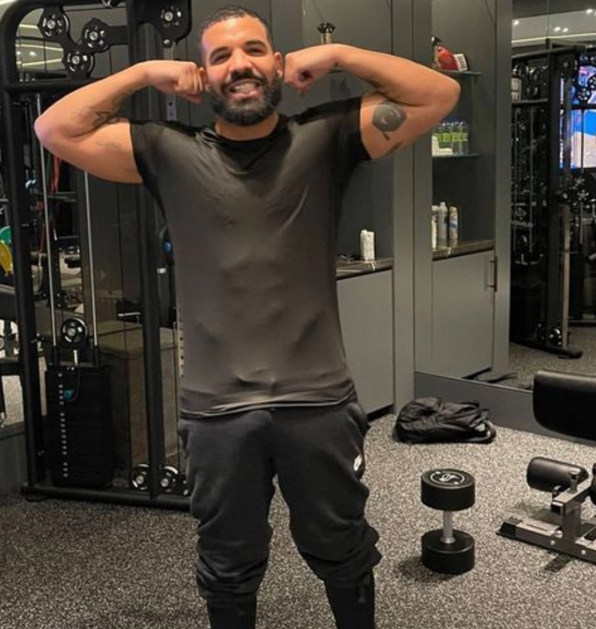 Drake flexing his arms in a gym while wearing a fitted black T-shirt, with weight machines and a dumbbell visible behind him