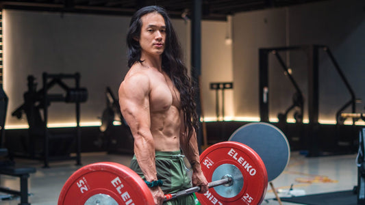 Eugene Teo holding a loaded barbell with red Eleiko plates in a gym, showing his muscular physique and long hair