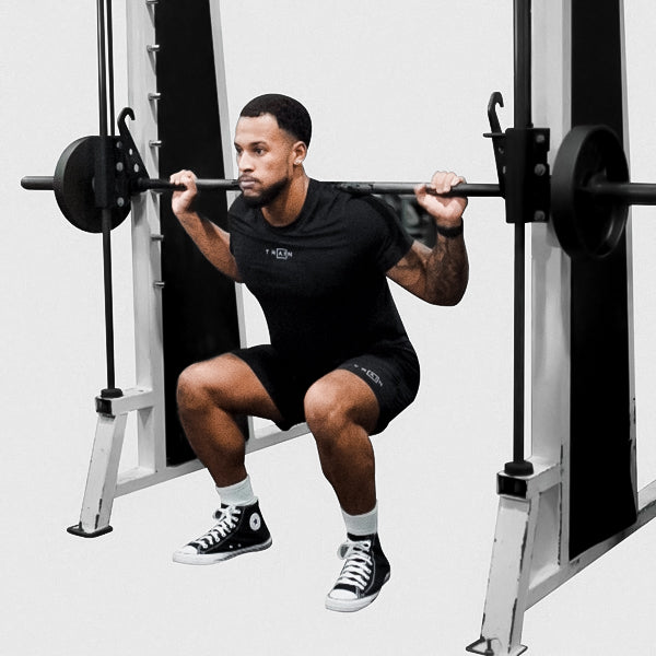 Athlete in black gym clothes performing a loaded Smith-machine squat against a light background