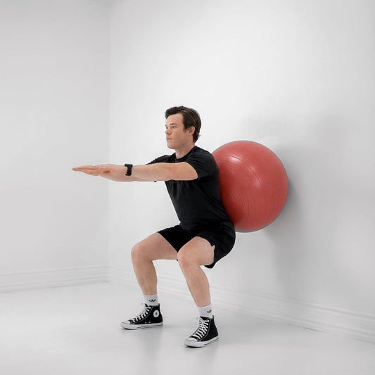 Man in black workout clothes performing a Swiss-ball wall squat with a red exercise ball against a white wall