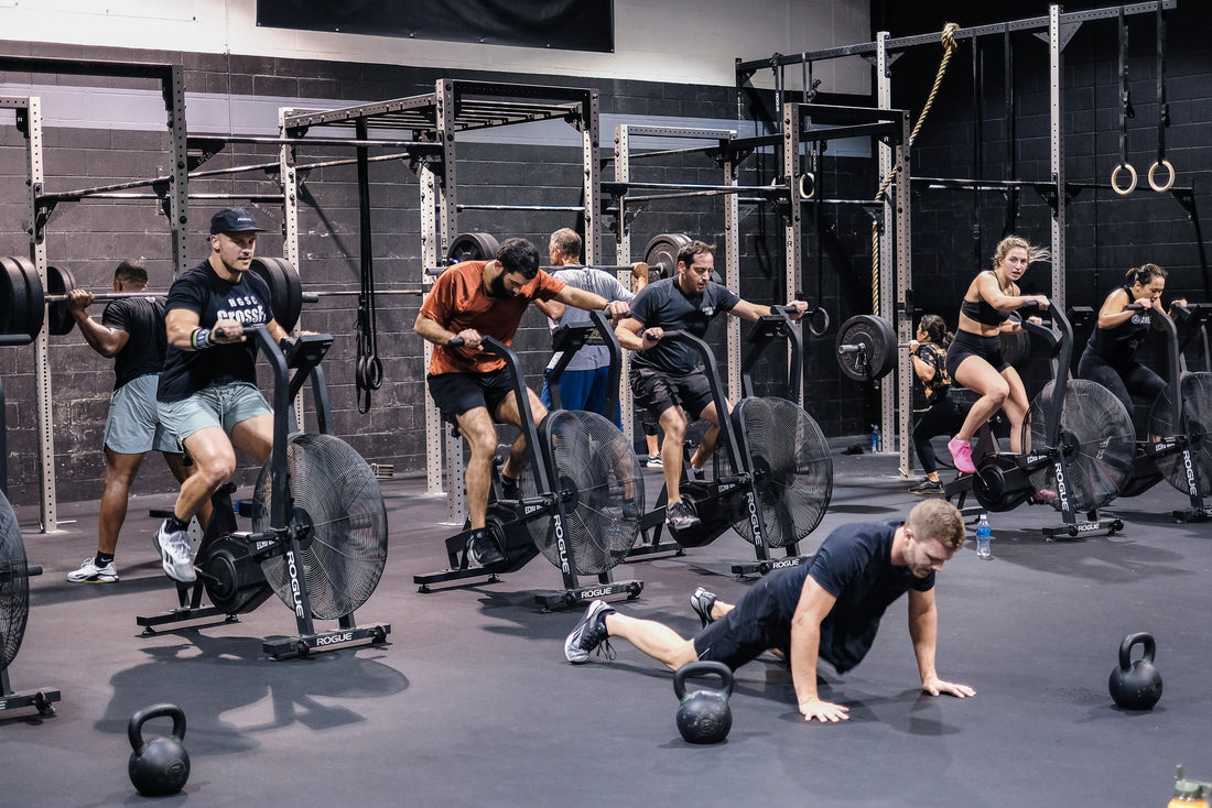 CrossFit gym group riding air bikes while another athlete works on the floor beside kettlebells