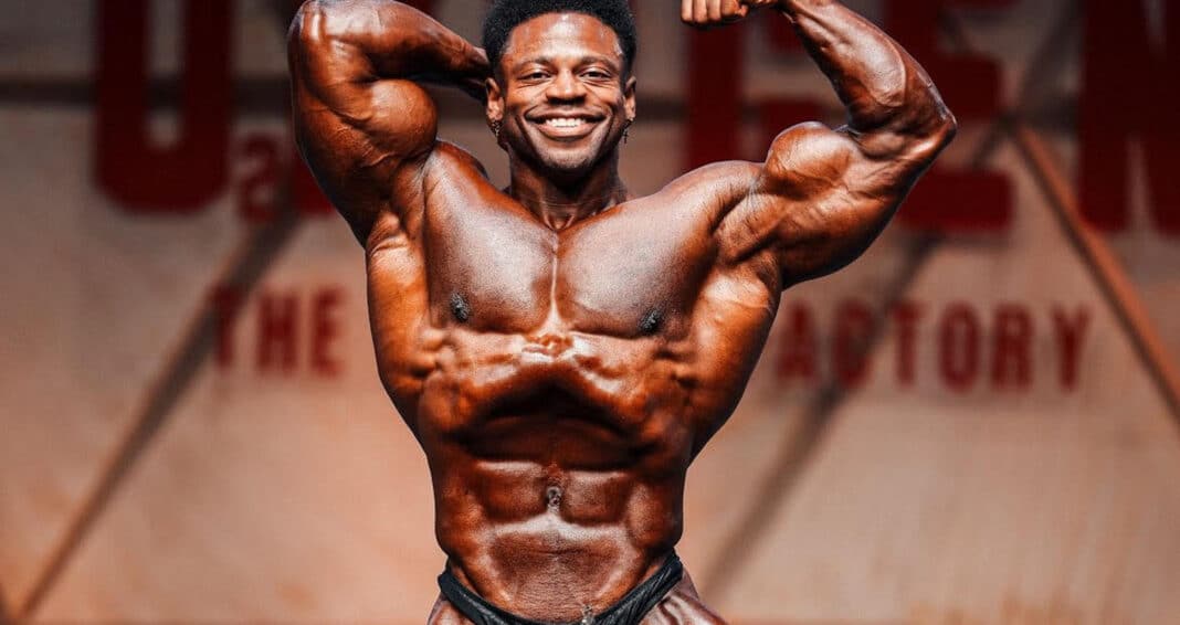 Breon Ansley smiling and flexing his arms in a front double-biceps pose onstage