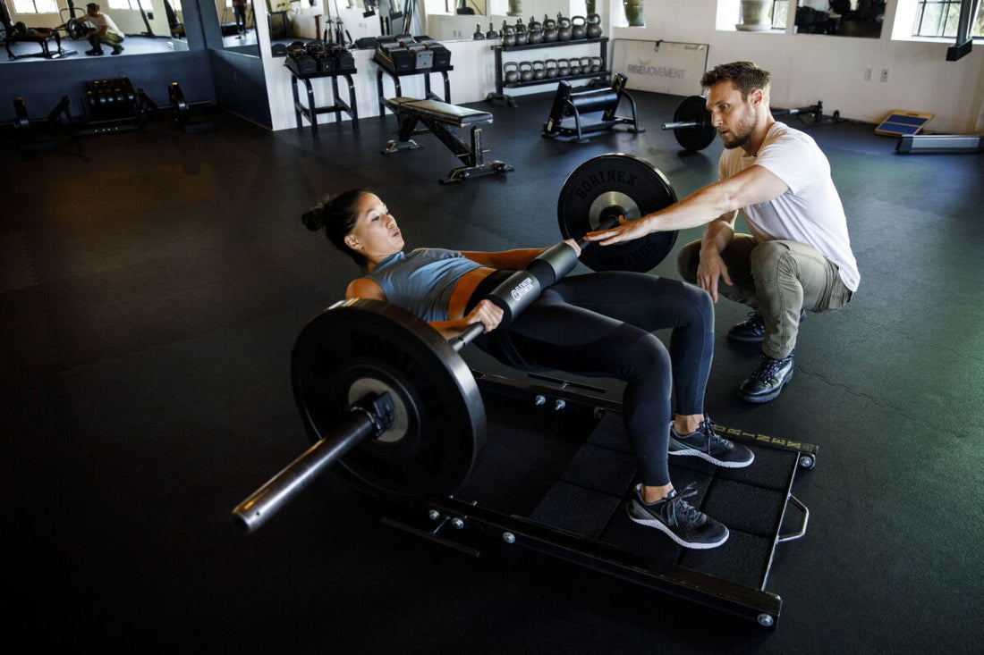 Brie Larson performing a heavy barbell hip thrust in a gym while trainer Jason Walsh watches her technique