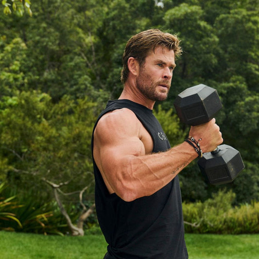 Chris Hemsworth holding dumbbells at shoulder height during an outdoor workout